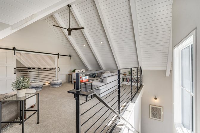Bright modern loft living area with vaulted white shiplap ceiling, black metal railing and ceiling fan, sliding barn doors, gray sectional, pouf seats, and large window letting in natural light.
