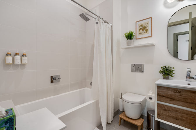 Bright, spa-like modern bathroom with white tiled tub and rain shower, wall soap dispensers, white shower curtain, wall-mounted toilet on a small wooden step, wood-veneer vanity with round mirror and potted plants.