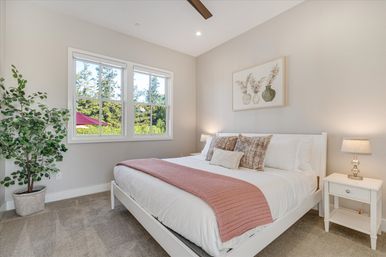 Sunlit cozy master bedroom with white bedframe, white bedding and a pink knit throw, textured pillows, bedside tables with lamps, potted plant by a large window overlooking trees and a red patio umbrella, neutral modern decor.