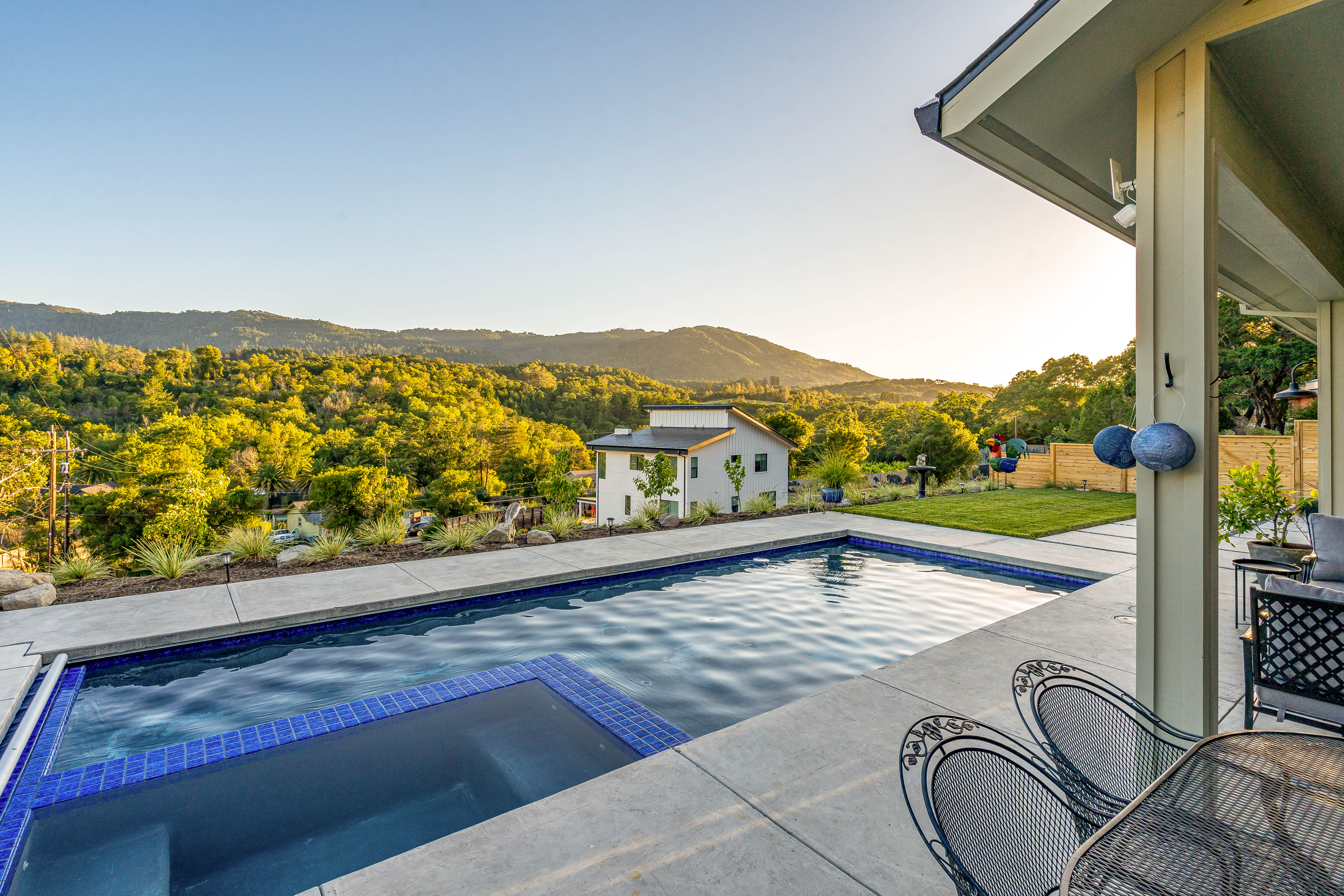 Rectangular backyard pool with attached blue-tiled spa on a concrete patio, covered seating and metal chairs in the foreground, modern white house and sunlit green hills and mountain view in the background — serene hillside outdoor living at sunset.