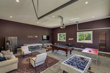 Finished garage game room with a pool table, foosball and ping-pong tables, air hockey, sofa seating on area rugs, stainless steel refrigerator, wall-mounted TV and neon sign, with windows looking out to a green yard.