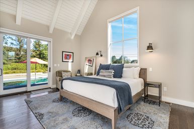 Sunny contemporary bedroom with vaulted white-beamed ceiling, wooden platform bed dressed in white linens and navy throw, patterned rug and bedside tables, large window and sliding glass doors opening to a backyard pool and umbrella.