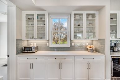 Sunlit white kitchen with glass-front cabinets, gray subway tile backsplash, central window overlooking trees and blue sky, countertop toaster and vase of daisies.