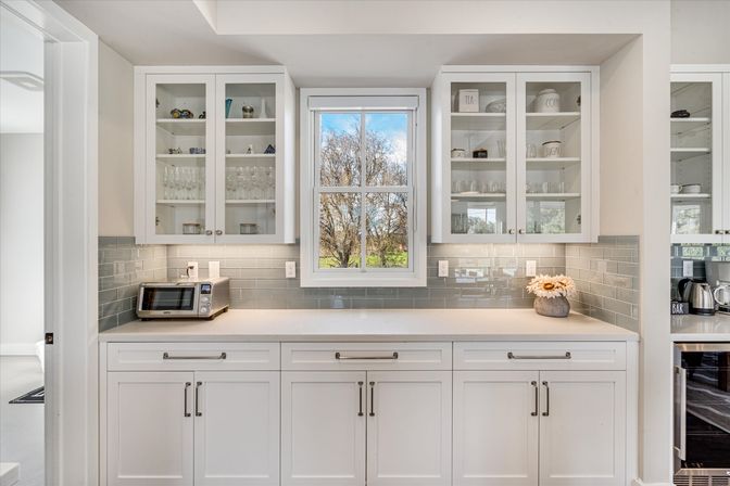 Sunlit white kitchen with glass-front cabinets, gray subway tile backsplash, central window overlooking trees and blue sky, countertop toaster and vase of daisies.