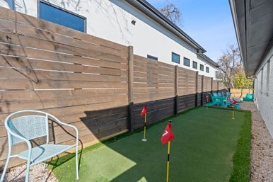 Narrow residential side yard featuring a mini putting green with red flags, horizontal wood privacy fence, light-blue metal chair in foreground and teal Adirondack chairs beside a modern white house with small windows.