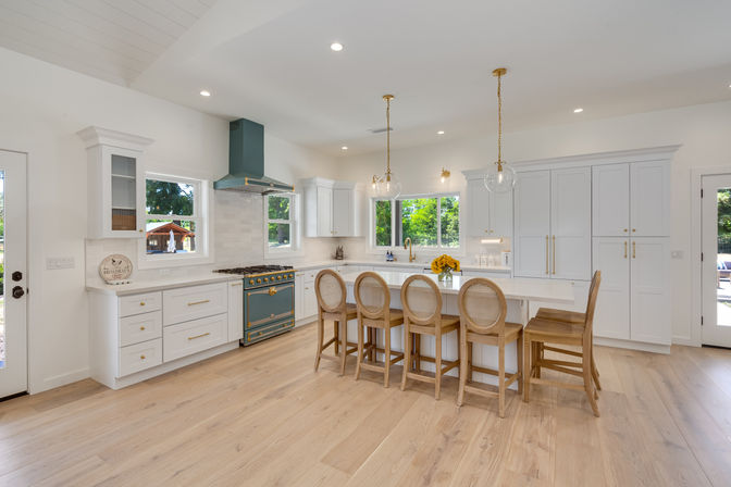 Bright open-concept modern kitchen with white cabinetry, large island breakfast bar with five wooden stools (four rattan-back), teal blue range and hood, brass pendant lights, light oak floors and sunlit windows
