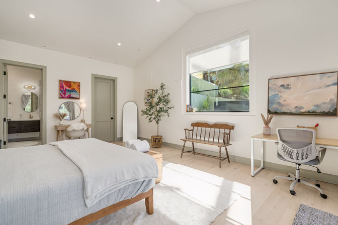 Bright modern bedroom with wooden bed, cozy white linens, vanity and round mirror, bench and desk with ergonomic chair by a large window overlooking a sunlit hillside, neutral Scandinavian decor and indoor plant.