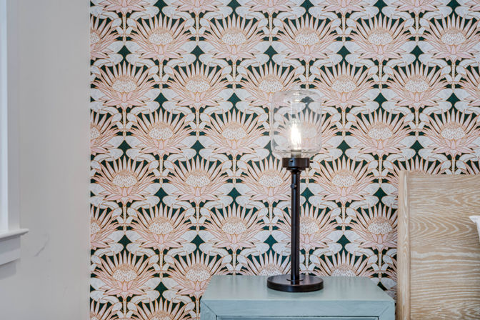 Lit glass-shade table lamp on a blue bedside table against pink and green repeating floral wallpaper, next to a light wood headboard — modern bedroom decor