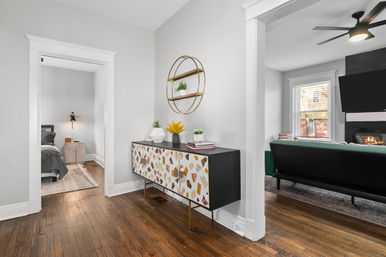 Bright modern home entryway with hardwood floors, patterned mid-century console table and round gold wall shelf with plants, view into bedroom and living room with green sofa, wall-mounted TV and fireplace