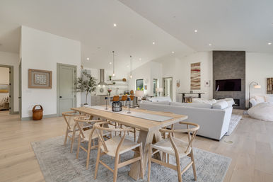 Sunlit open-concept living, dining and kitchen with a light wood farmhouse table and wishbone chairs, neutral sofa, pendant lights and a gray stone fireplace