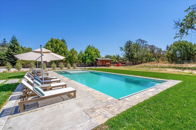 Sunny rural backyard with a rectangular turquoise pool, stone deck lined with cushioned lounge chairs and a large umbrella, green lawn, trees, and a wooden shed in the background.