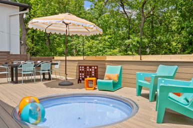 Sunny backyard deck patio with round inset plunge pool and colorful beach balls, striped umbrella, turquoise lounge chairs, outdoor dining set and leafy green treeline backdrop