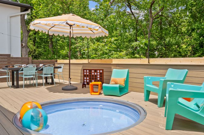 Sunny backyard deck patio with round inset plunge pool and colorful beach balls, striped umbrella, turquoise lounge chairs, outdoor dining set and leafy green treeline backdrop