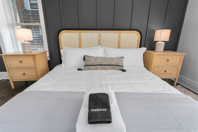 Stylish guest bedroom interior with rattan-accent headboard, dark gray paneled accent wall, crisp white linens and patterned lumbar pillow, matching light-wood nightstands with lamps, and folded towels (one labeled “makeup”) at the foot of the bed.