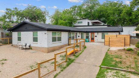 Modern single-story white brick home with black trim and a cheerful orange front door, gravel front yard, wooden fence, concrete driveway, patio chairs, and mature trees in a quiet suburban setting