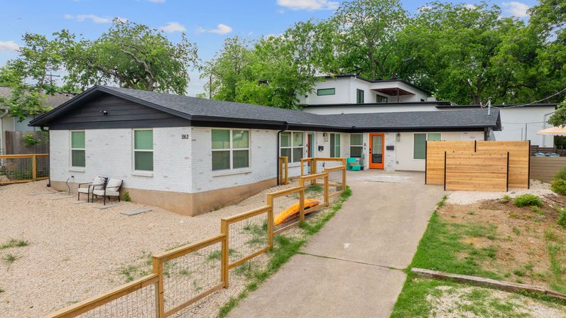 Modern single-story white brick home with black trim and a cheerful orange front door, gravel front yard, wooden fence, concrete driveway, patio chairs, and mature trees in a quiet suburban setting