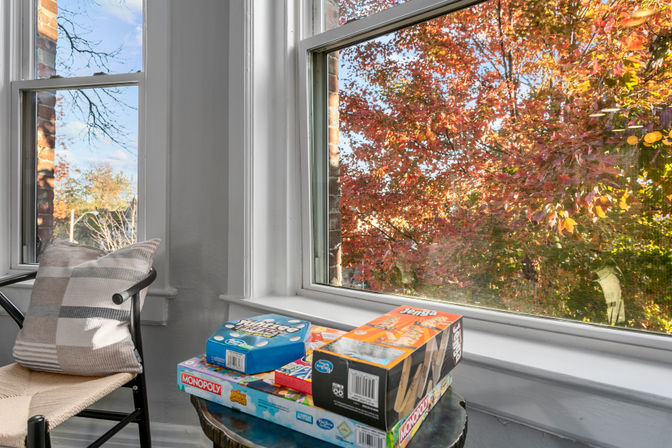 Cozy home window nook with a cushioned chair and a stack of board games on a small table, large windows framing bright orange-red autumn leaves outside.