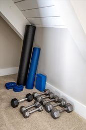 Home workout corner under a sloped white ceiling with a black foam roller, blue rolled yoga mat and block, blue ankle weights, and several hex dumbbells on beige carpet.