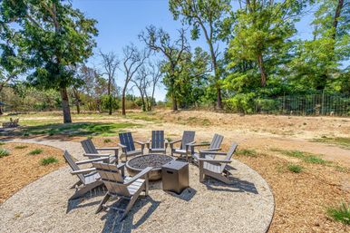 Sunny outdoor seating area with eight gray Adirondack chairs arranged around a circular fire pit on a gravel patio in a park-like backyard with mature trees and dry grass.