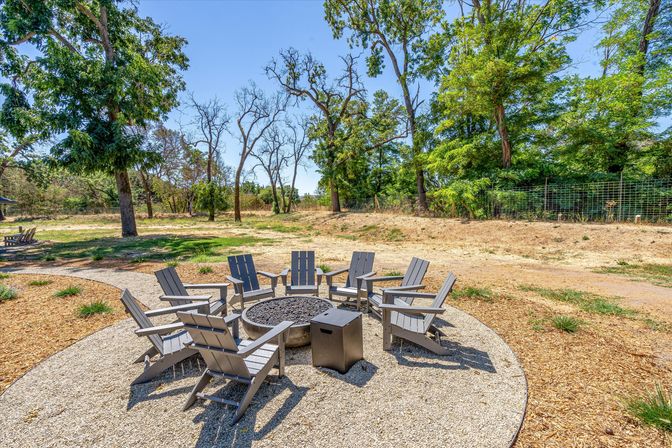 Sunny outdoor seating area with eight gray Adirondack chairs arranged around a circular fire pit on a gravel patio in a park-like backyard with mature trees and dry grass.