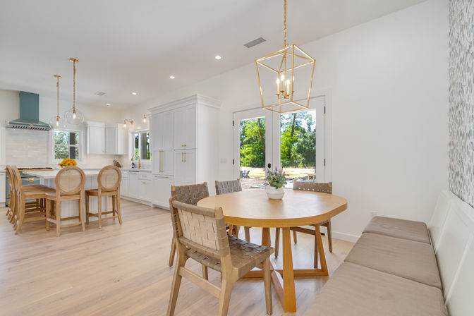 Bright modern open-plan kitchen and dining area with round oak table and woven chairs, white cabinetry, island with cane-back stools, gold pendant lights, and glass doors opening to a green backyard.