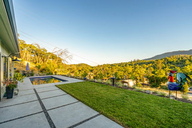 Hillside backyard at golden hour with a rectangular plunge pool, modern concrete paver patio, manicured lawn and colorful rooster garden sculpture overlooking a sunlit valley and tree-covered hills