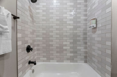 Clean modern bathtub-shower with light gray subway tiles, matte black showerhead and faucet, wall-mounted soap dispenser and folded white towel on a towel bar.