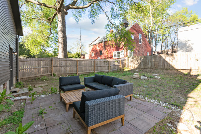 Sunny suburban backyard patio with modern wicker outdoor seating and wooden coffee table on stone pavers, fenced yard with large shade tree and a red house beyond.