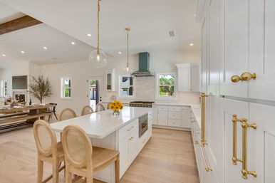 Sunlit open-plan modern farmhouse kitchen with large white island, brass hardware, light wood floors, rattan-back stools, teal range hood, globe pendant lights, and adjacent dining area