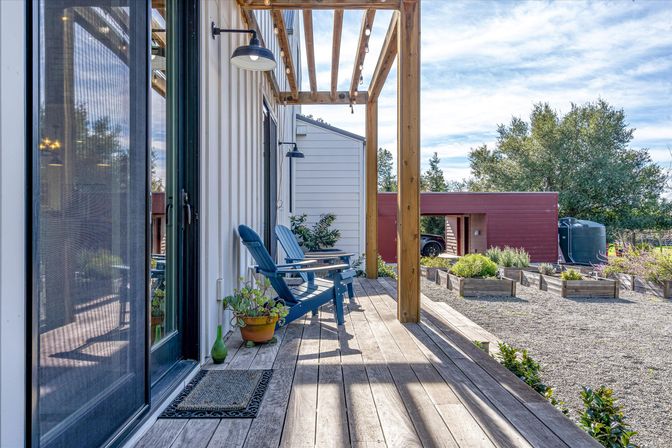 Sunlit wooden deck with pergola, blue Adirondack chairs, potted plants, gravel yard and raised garden beds beside a red outbuilding
