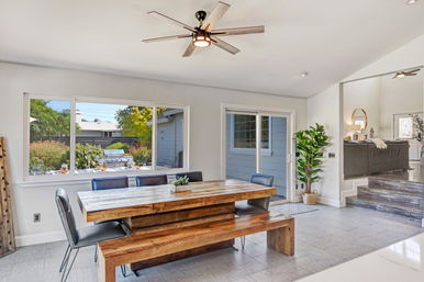 Bright, open-concept suburban dining area with a rustic wooden table and bench, black chairs, modern ceiling fan, large windows and sliding glass door overlooking a backyard patio with a grill, and a tall potted plant.