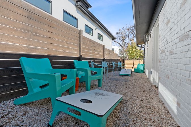 Narrow side-yard outdoor patio with turquoise plastic lounge chairs and matching cornhole boards on gravel, bordered by a wooden privacy fence and white brick wall under a clear blue sky.