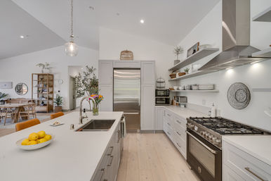 Bright open-concept modern kitchen with a large white island, stainless-steel refrigerator and gas range, pendant light, open shelves with dishware, vase of flowers and bowl of lemons, dining area in background