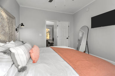 Contemporary bedroom interior with white bedding, coral throw and accent pillow, carved wooden headboard, black wall sconce, leaning full-length mirror, wall-mounted TV, and neutral gray walls.