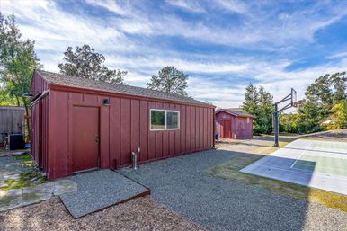 Sunny suburban backyard with two red wooden storage sheds, gravel yard, and a half basketball court under a bright blue sky with wispy clouds.