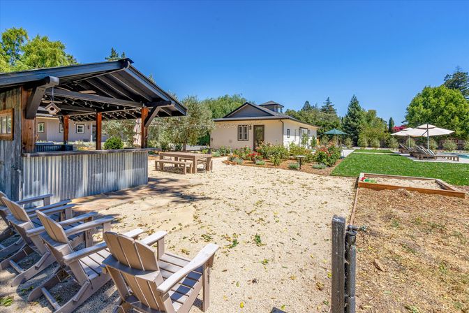 Sunny backyard oasis with a covered corrugated-metal outdoor bar and string lights, Adirondack chairs on a gravel lounge area, picnic table, landscaped garden beds, a pool with umbrellas and loungers, and a small guest cottage.