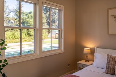 Cozy neutral bedroom with white bed and bedside lamp, large windows framing a sunny backyard pool and leafy garden view.