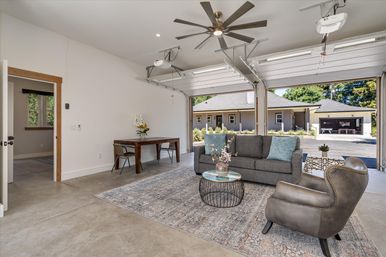 Bright modern garage living space with concrete floor and large ceiling fan, gray sofa and leather armchair on a patterned rug, round wire coffee table, wooden desk, and open overhead doors opening to a suburban driveway and neighboring houses.