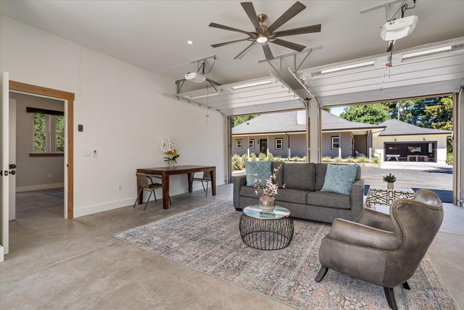 Bright modern garage living space with concrete floor and large ceiling fan, gray sofa and leather armchair on a patterned rug, round wire coffee table, wooden desk, and open overhead doors opening to a suburban driveway and neighboring houses.