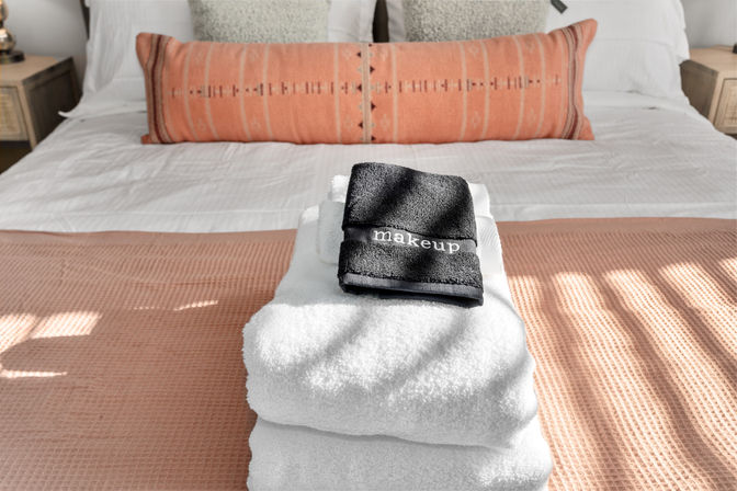 Stacked white bath towels topped with a small black towel labeled 'makeup' on a blush textured throw at the foot of a sunlit modern guest bedroom bed with a coral lumbar pillow.