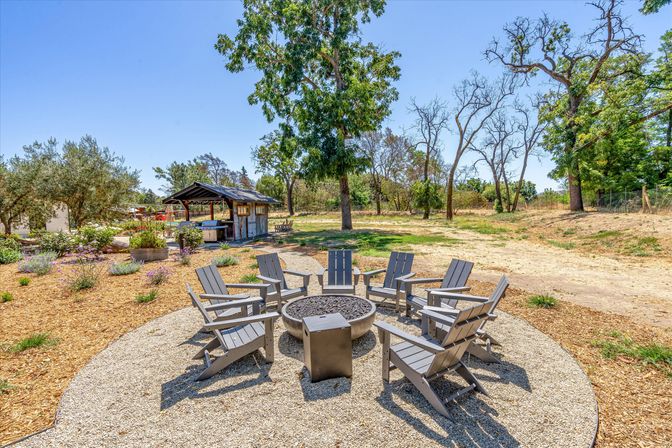 Outdoor fire pit seating: circular gravel patio with eight gray Adirondack chairs around a central fire bowl, set in a sunny rustic backyard garden with trees, wildflowers and a small wooden pavilion