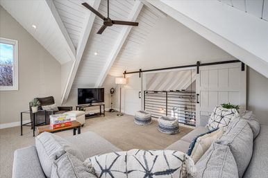 Bright cozy attic loft living room with vaulted white beamed ceiling, ceiling fan, gray sectional, TV, sliding barn-style doors and open railing overlooking lower level