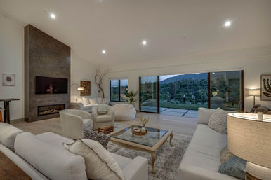 Cozy modern open-concept living room with neutral sofas, armchairs and glass-top coffee table, wall-mounted TV above a sleek fireplace, and wide sliding glass doors opening to rolling hills and a mountain view.