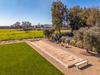 Aerial view of a sandy bocce court in a rural backyard, bordered by a manicured green lawn, gravel seating area with wooden benches and Adirondack chairs, olive trees and open pasture under a clear blue sky.