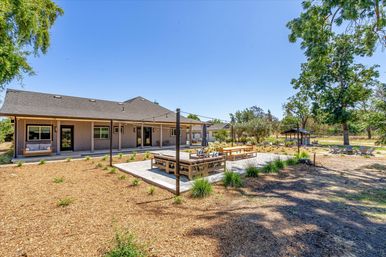 Spacious suburban backyard with a single-story house, concrete patio featuring wooden lounge and dining benches, string lights, landscaped mulch beds, gazebo and fire‑pit seating under trees on a sunny day.