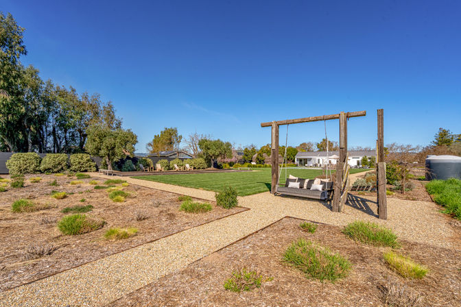 Spacious landscaped backyard with gravel pathways, manicured lawn, timber-framed hanging bench with cushions, low shrubs and mature trees under a clear blue sky with a house in the background.