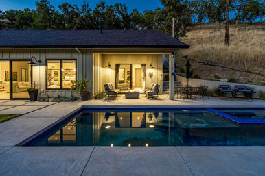 Twilight view of a modern hillside home backyard with a lit rectangular pool reflecting patio lights, cozy covered seating area with a fire pit, outdoor dining table and lounge chairs on a concrete deck.