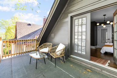 Cozy second-floor balcony patio with two wicker chairs and a glass-top table by a French sliding door leading into a bedroom, overlooking neighboring rooftops and leafy trees.