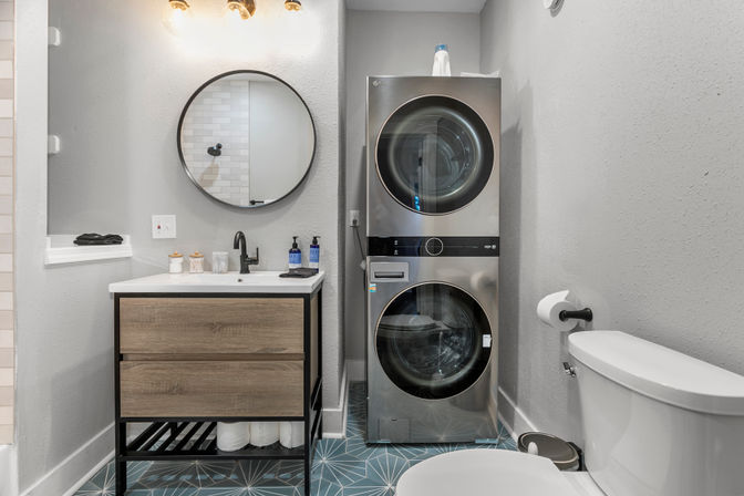 Compact modern bathroom with stacked stainless-steel washer and dryer, wood vanity under round mirror, black faucet, blue geometric floor tile, gray walls and white toilet.