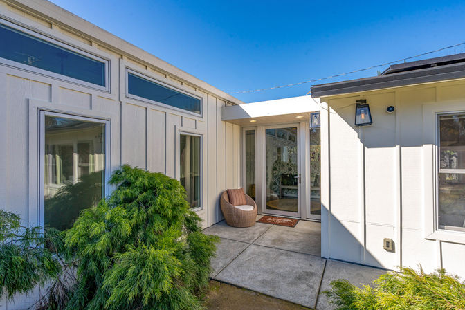 Sunny residential front entry courtyard of a white house with glass double doors and transom windows, a rattan lounge chair on a concrete patio and lush green shrubs under a bright blue sky.